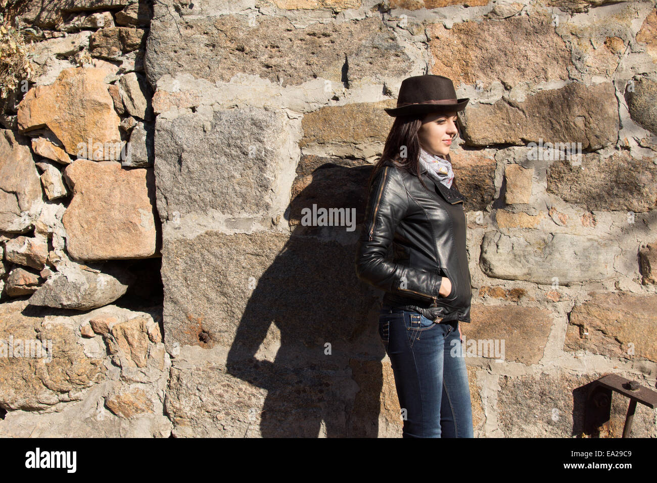 Stylish young woman in a black leather jacket, trendy hat and scarf standing sideways against an old stone wall in the sunshine Stock Photo