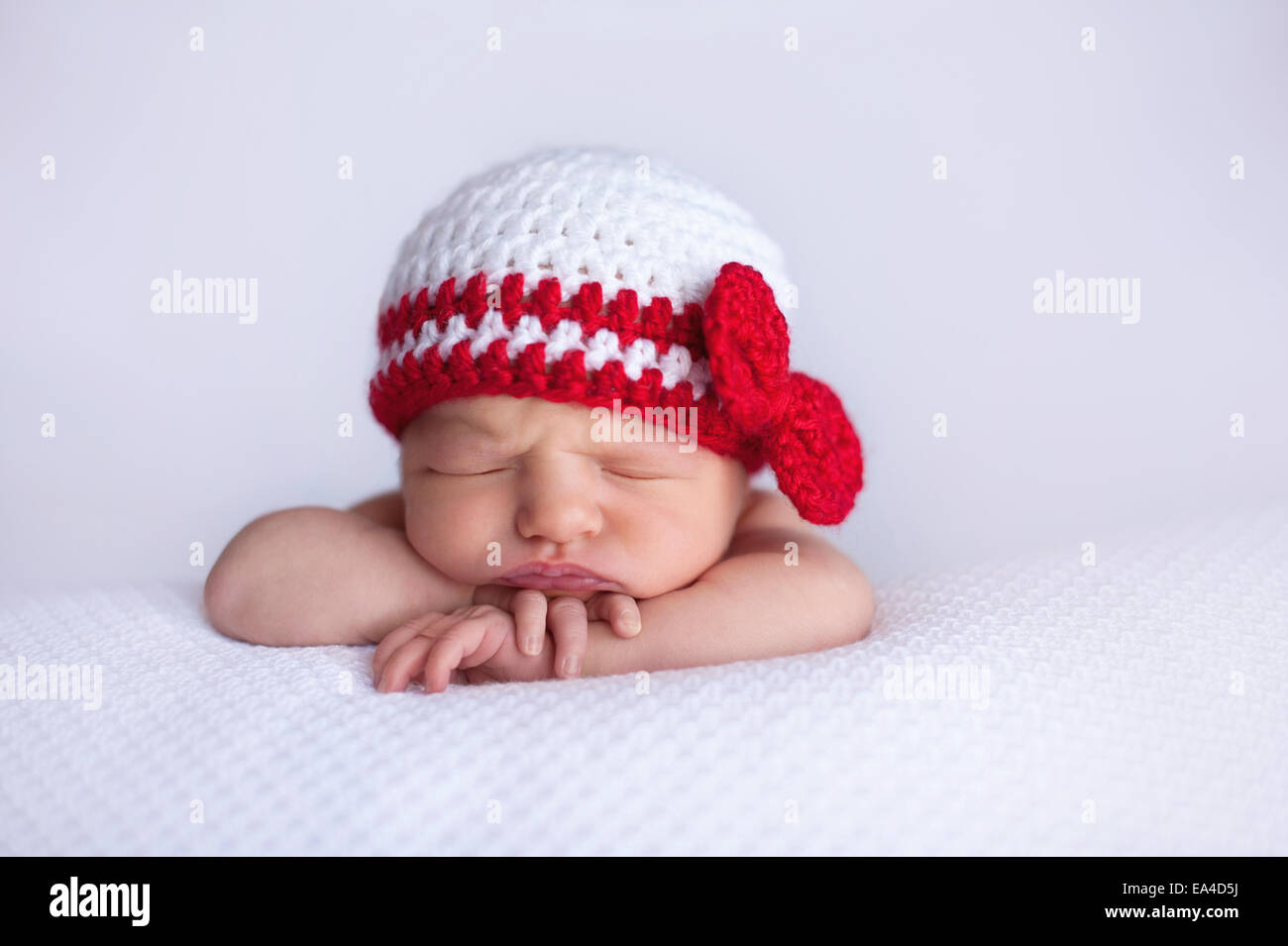 Portrait of a seven day old newborn baby girl. She is wearing a white and red crocheted hat and is sleeping on white, textured m Stock Photo