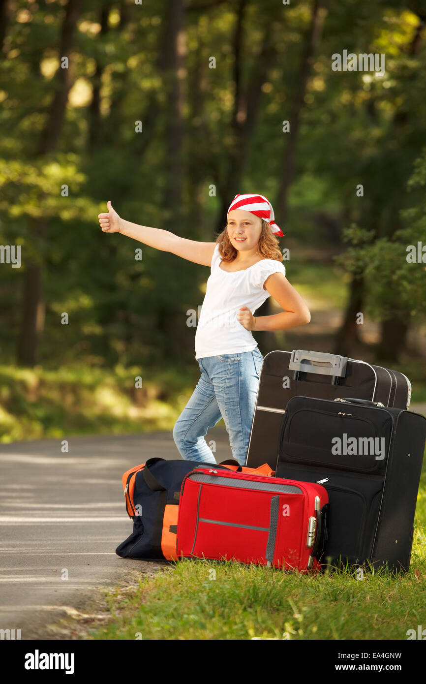 Young hitch-hiker girl standing on road side afternoon in forest with bags Stock Photo