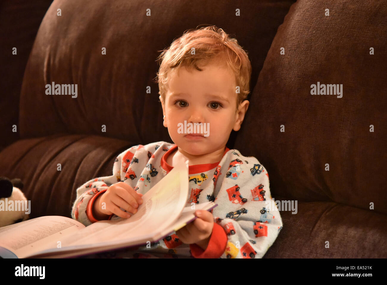 A young boy (two and a half years old) in pyjamas sat on a sofa reading a book just before bedtime Stock Photo