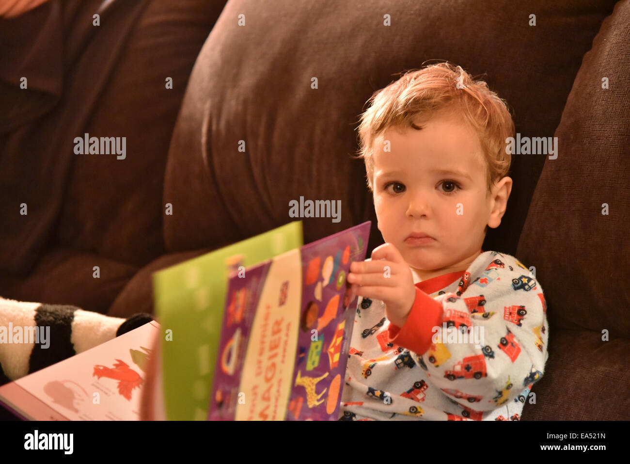 A young boy (two and a half years old) in pyjamas sat on a sofa reading a book just before bedtime Stock Photo