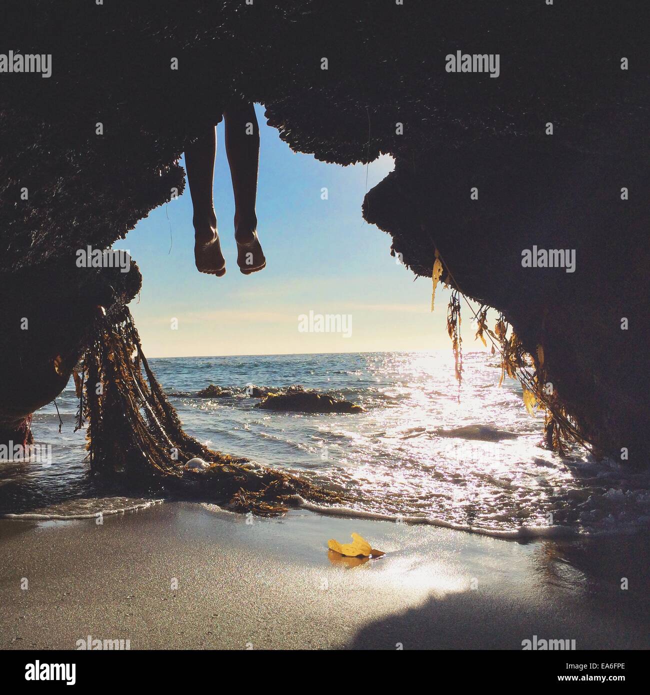 Person's legs hanging in front of cave entrance on beach Stock Photo