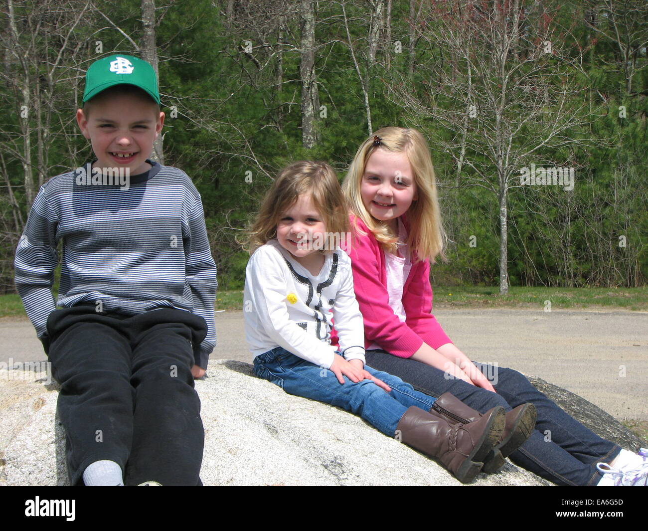 Three smiling children (8-9, 2-3, 6-7) sitting on boulder Stock Photo