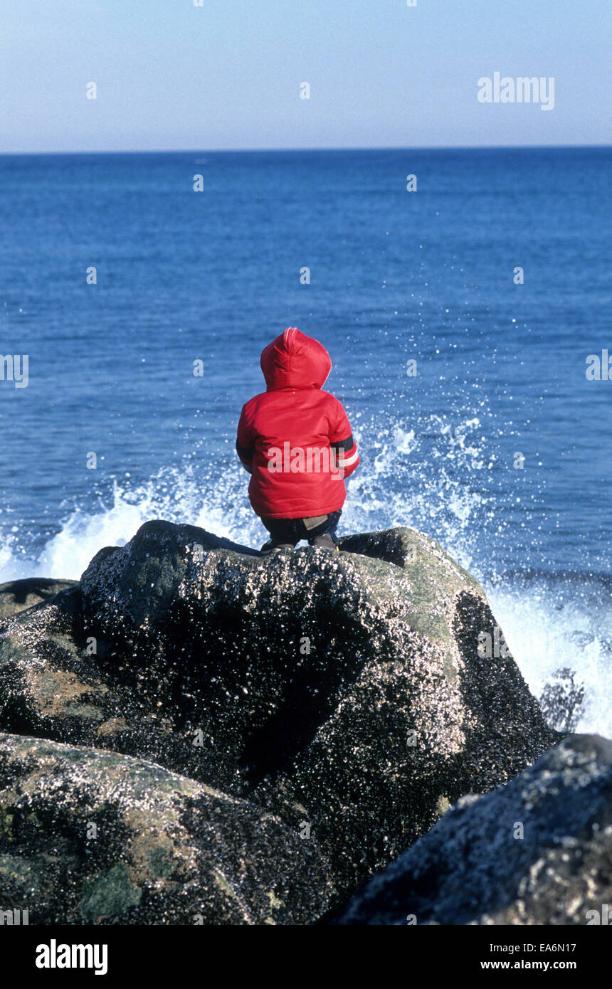 One child sitting on rocks by the ocean watching waves break. Stock Photo