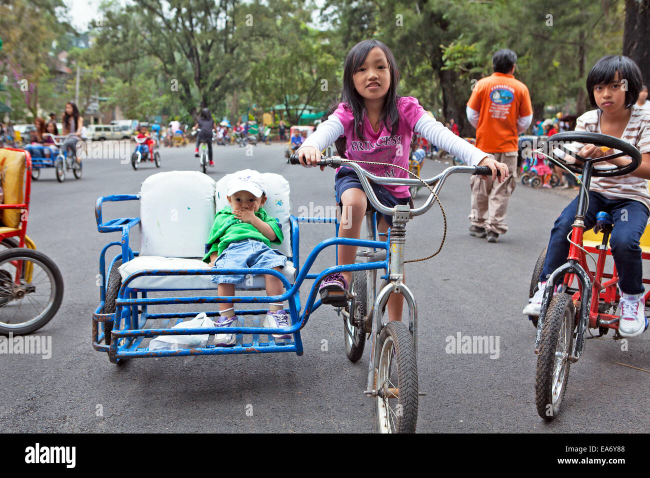 Filipino children riding bicycles in Burnham Park, Baguio City, Philippines. One young girl rides bike with sidecar with brother Stock Photo