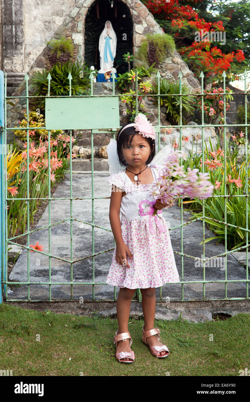 Cute young Filipino girl carries flowers and is dressed in pink for Sunday mass. Stands in front of garden gate. Stock Photo