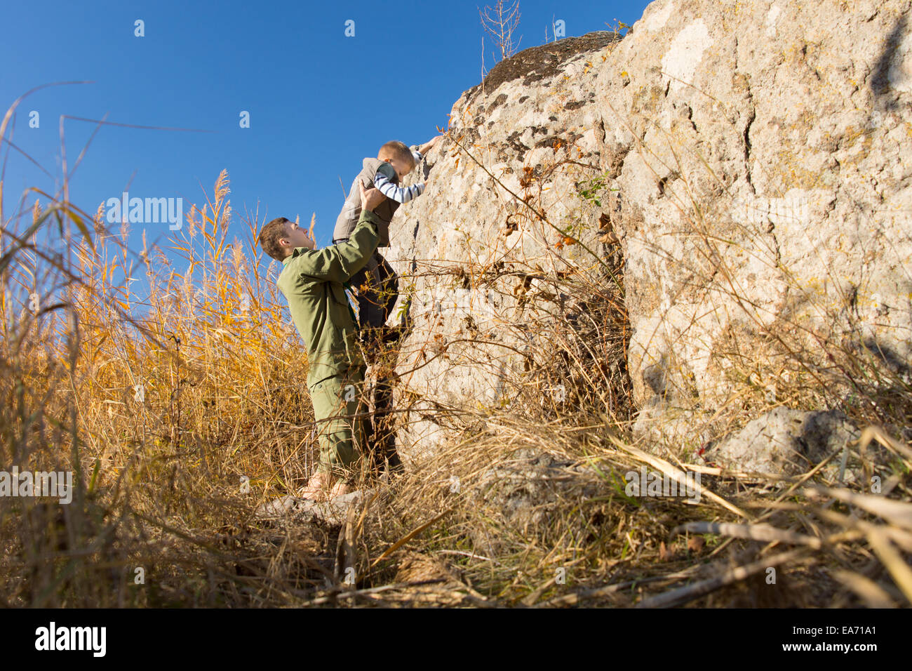 Man helping a young boy climb a rock supporting him around the waist with his hands as he finds a foothold Stock Photo