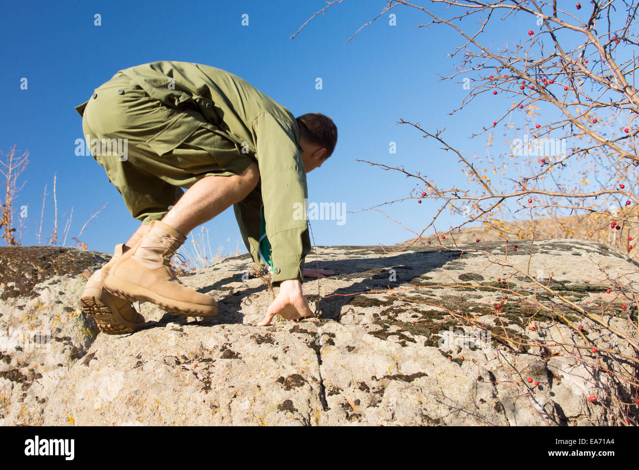 Side View of a Young Male Scout Climbing a Big Rock at the Field on a Tropical Climate with Light Blue Sky Background. Stock Photo