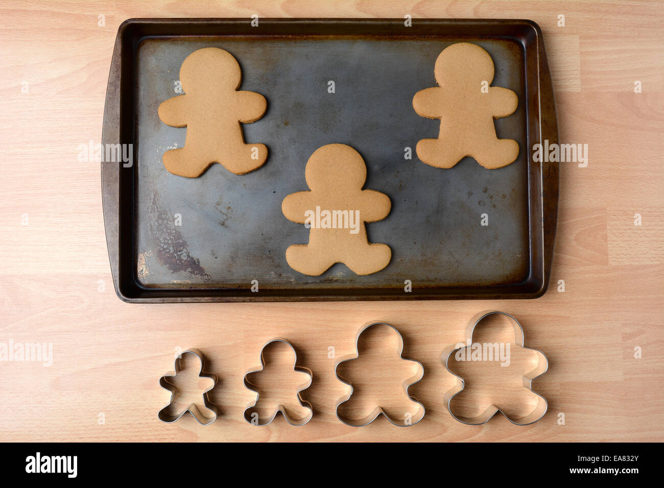 High angle shot of a cookie sheet with three gingerbread man cookies. Below the pan is a row of four cookie cutters of different Stock Photo