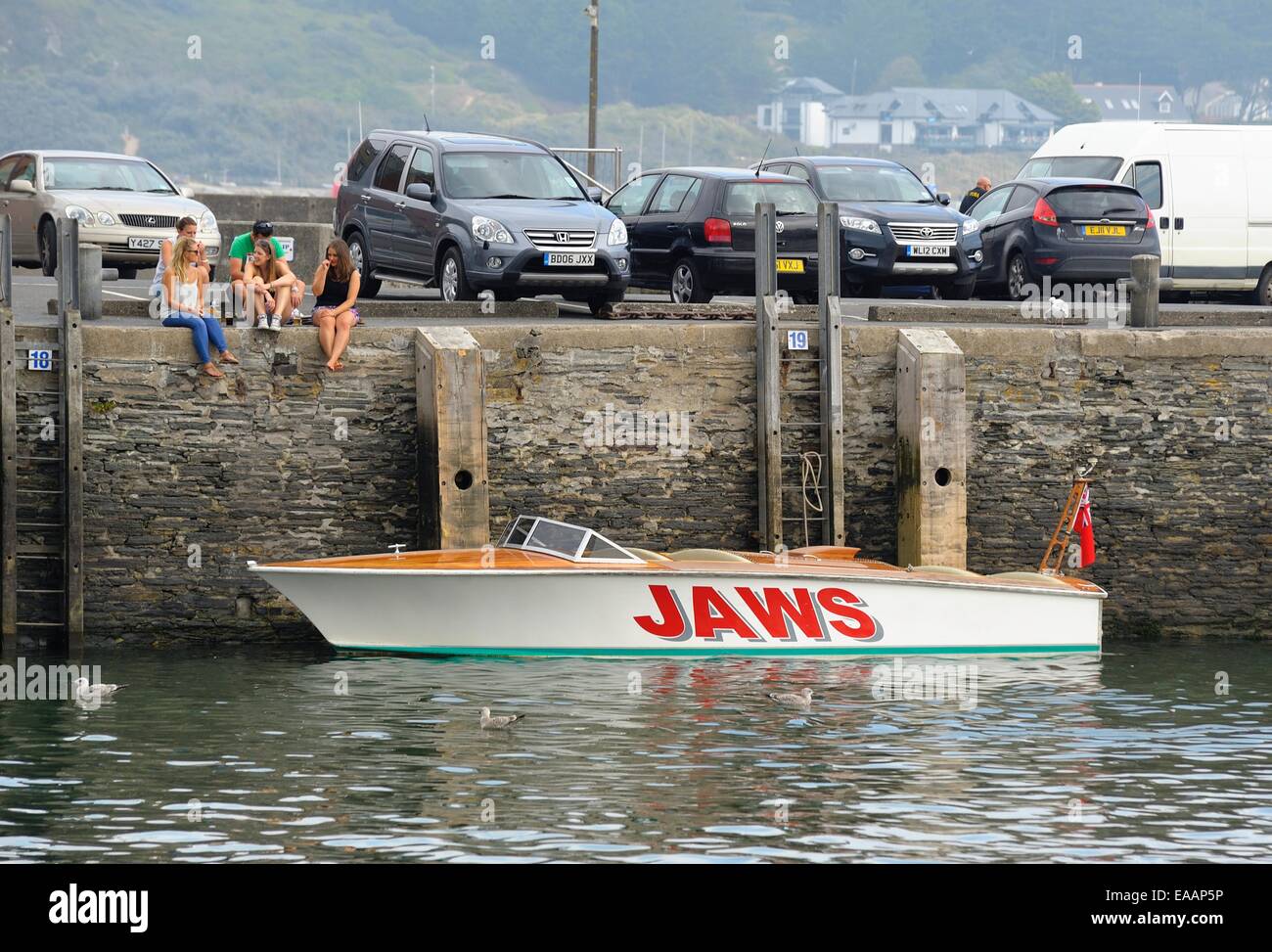 A group of teenagers sitting on the harbour wall next to a speedboat called Jaws.Padstow, Cornwall ,England ,uk Stock Photo
