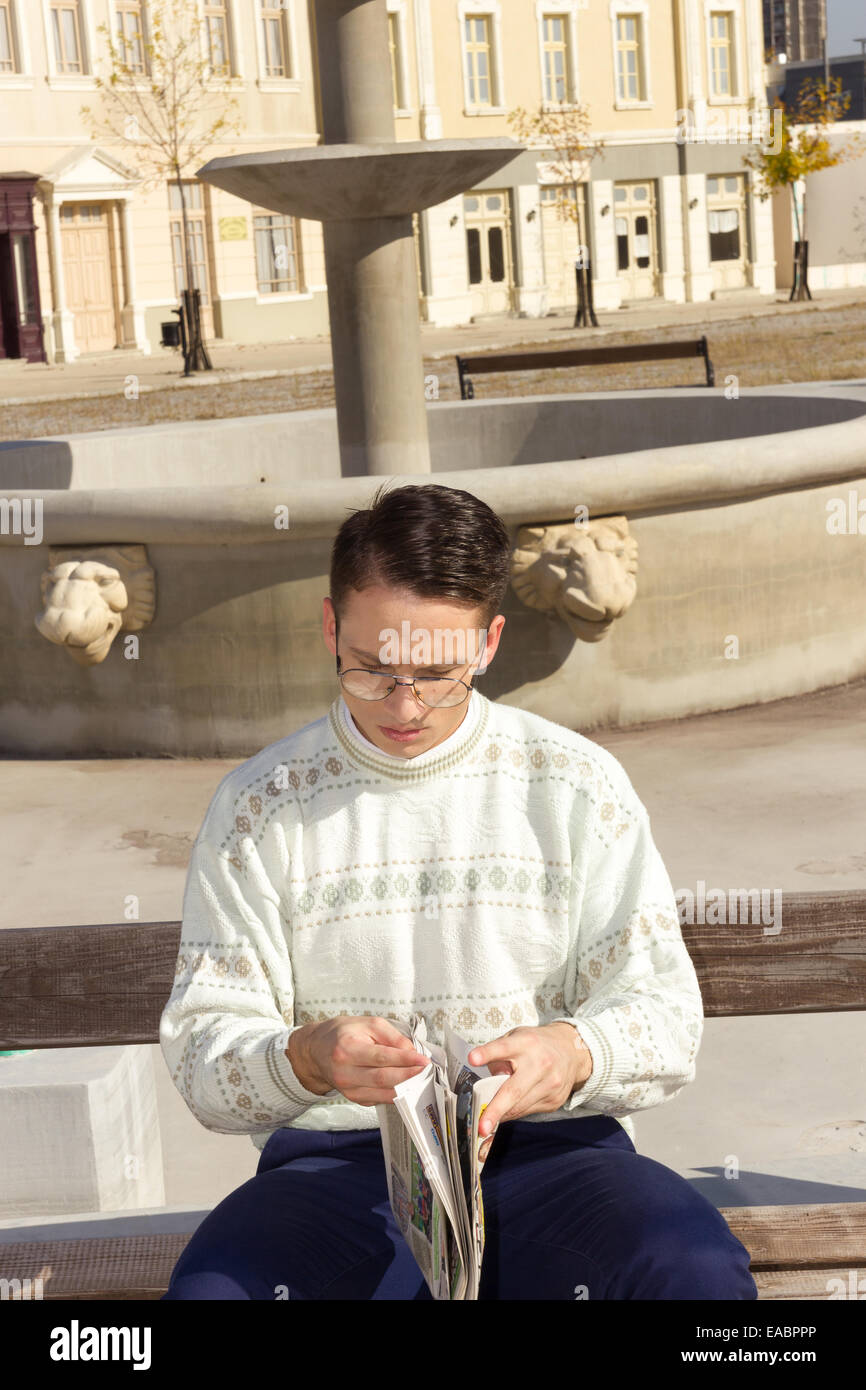 man with glasses in white sweater reading newspaper on bench next to fountain in town Stock Photo