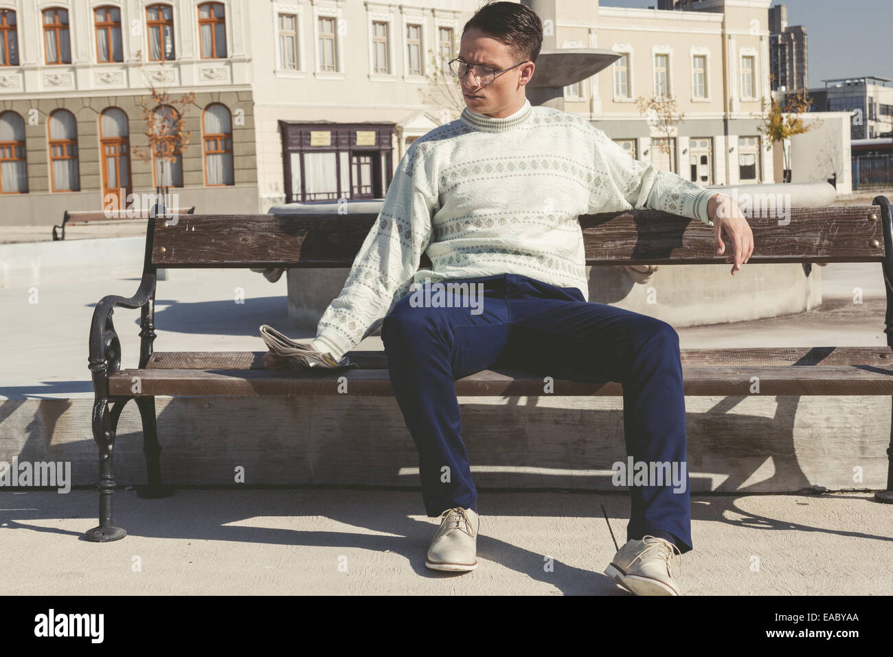 vintage photo of man with glasses in white sweater reading newspaper on bench next to fountain in town Stock Photo