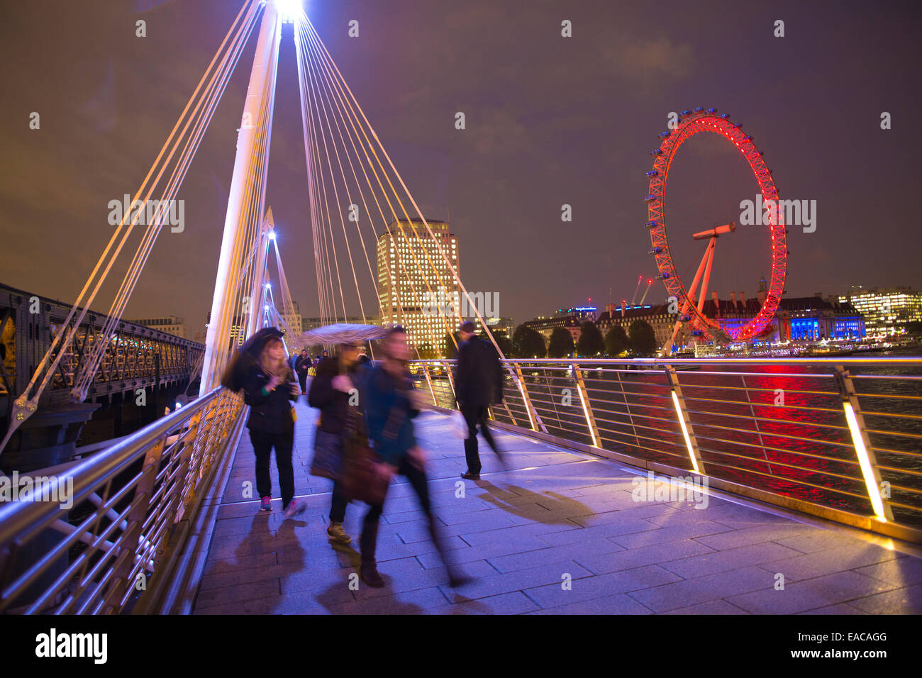 Illuminated Golden Jubilee Bridges run across the River Thames in London with the London Eye illuminated in red in the distance. Stock Photo