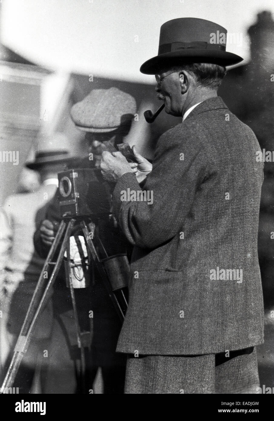 1930s, Historical picture showing a gentleman photographer wearing a suit, a hat and pipe, with his assistant standing by the camera which is on a tripod. The photographer is looking at two images in his hands, which are possibly glass negatives, Stock Photo
