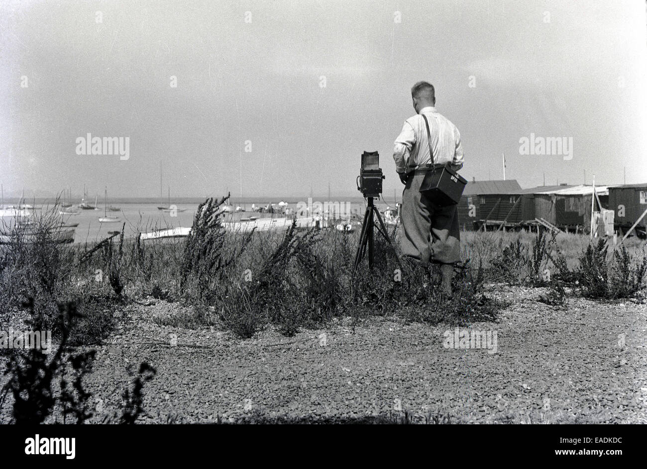 1930s, Historical picture of a male photographer using a large film box camera of the era on a wooden tripod to take a picture of the Norfolk coastline. Stock Photo
