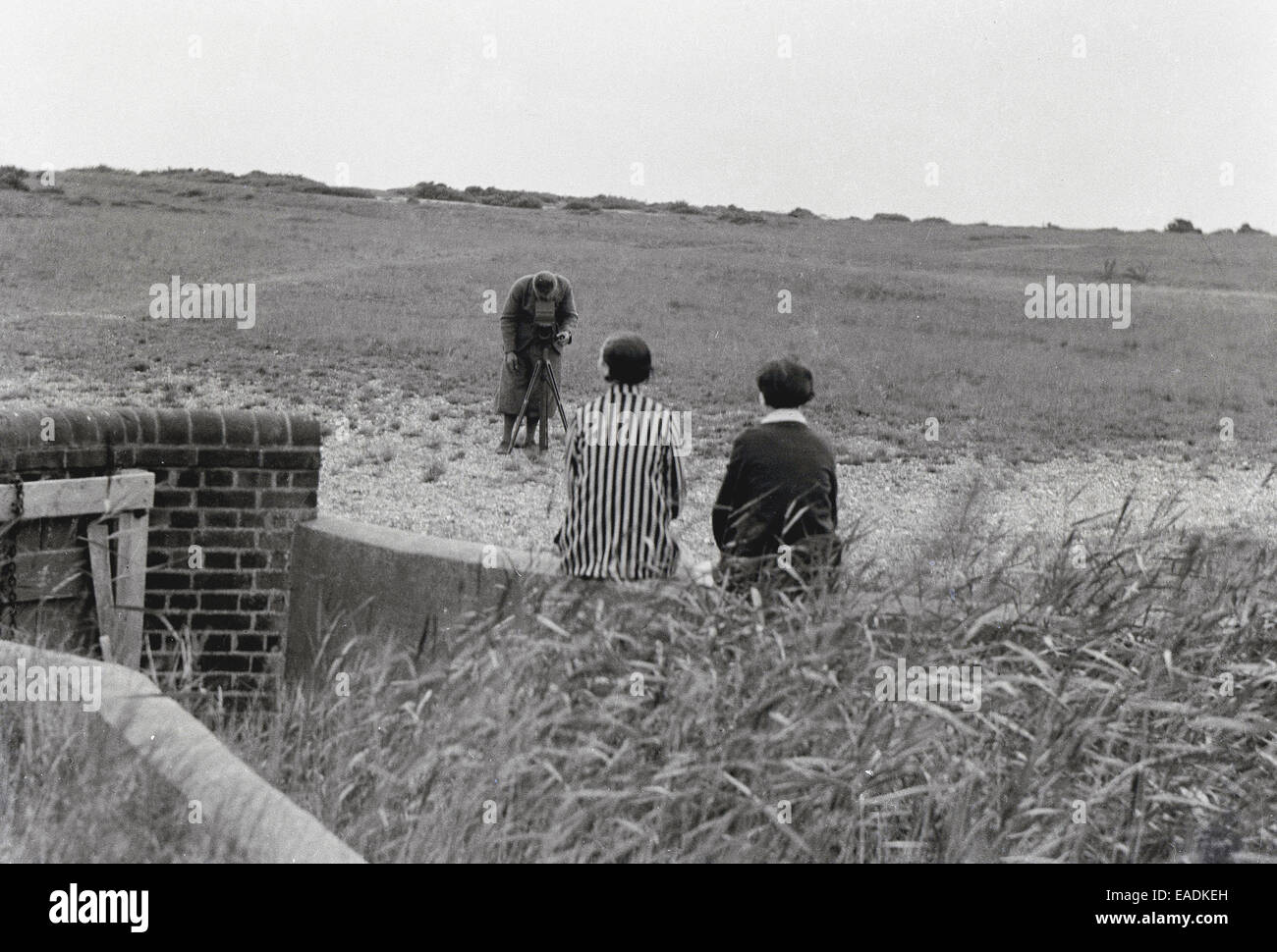 1930s, Historical picture showing a photographer in plus-fours using a large film camera of the era on a wooden tripod to take a picture of two ladies sitting outside on a wall at the Norfolk coast, England, UK. Stock Photo