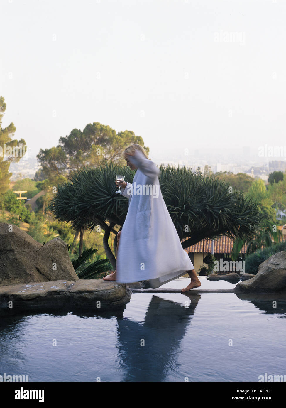 woman in white terry clothe robe walking across a swimming pool with a glass of wine in hand. Stock Photo