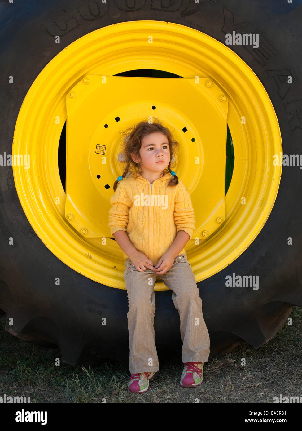 Young girl sitting on large yellow truck wheel and staring away pensively Stock Photo