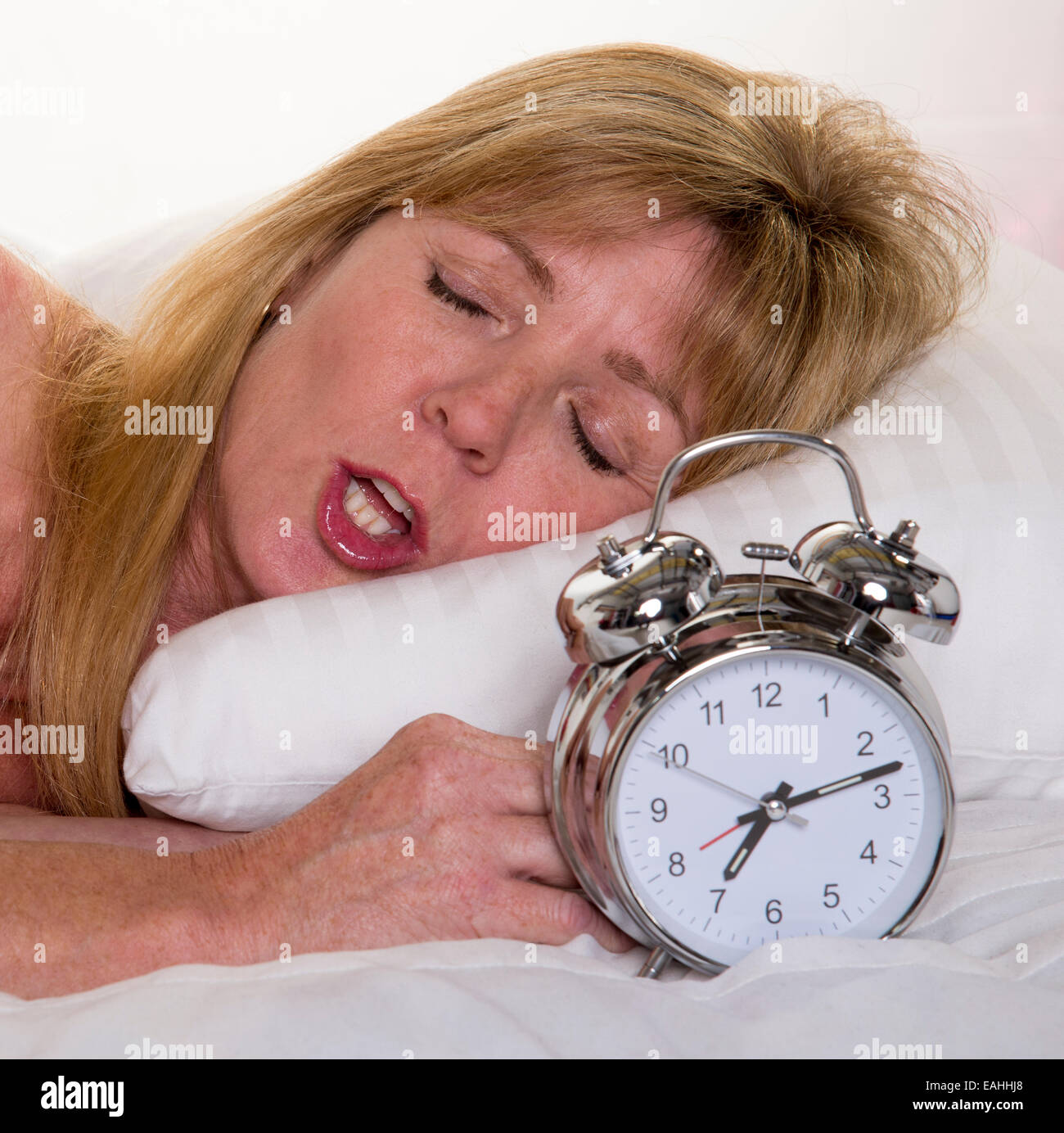 Early morning woman snoring in bed holding an alarm clock Stock Photo