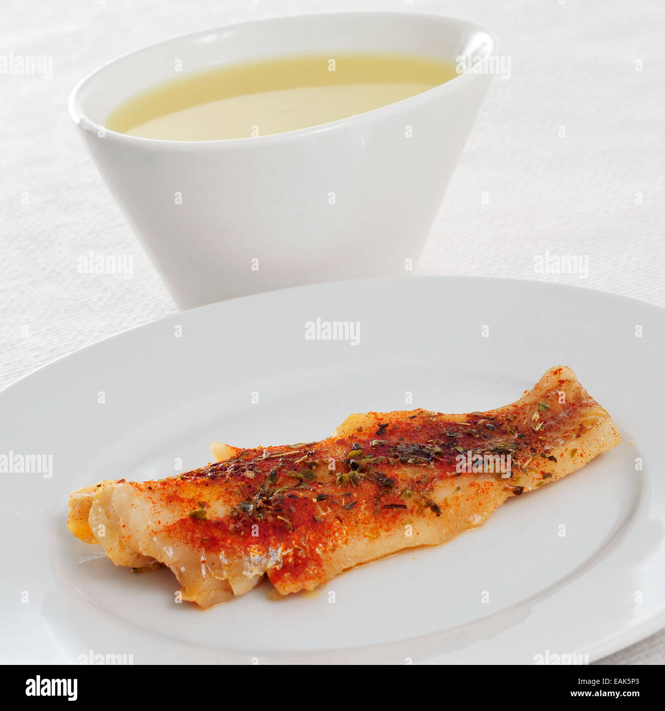 a bowl with consomme and a plate with a grilled hake fillet on a set table Stock Photo