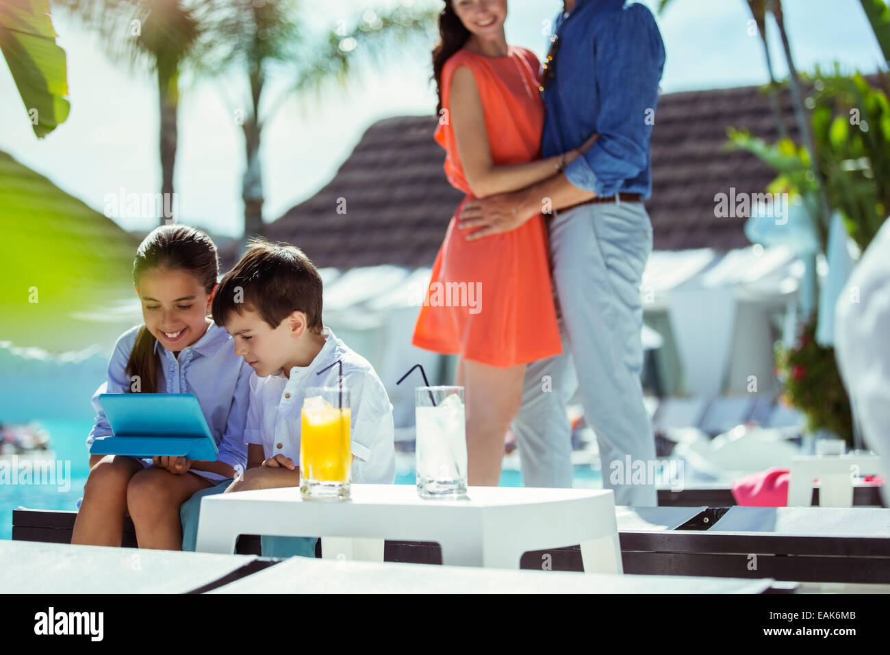 Brother and sister using digital tablet by resort swimming pool, parents embracing in background Stock Photo