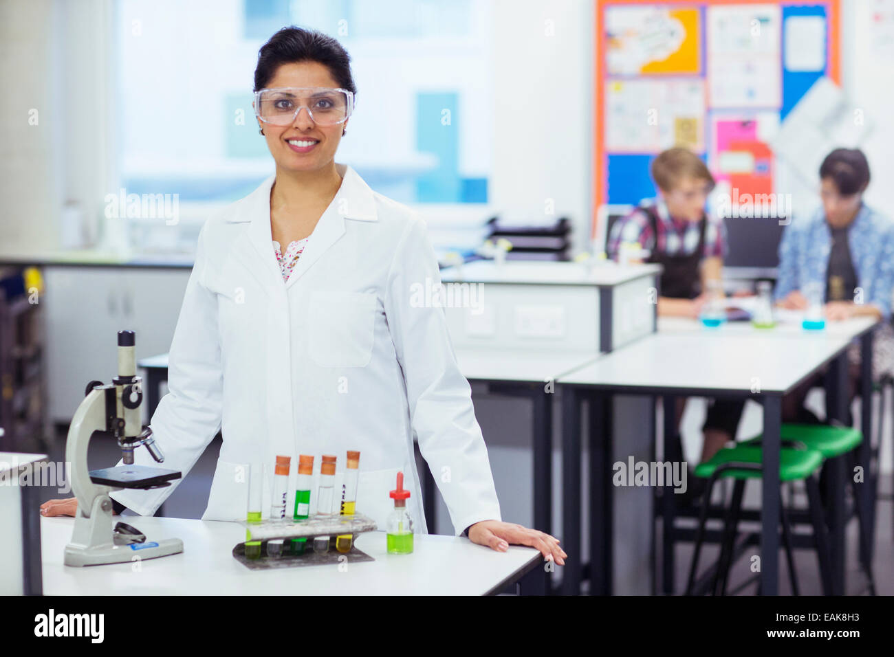 Portrait of smiling female teacher wearing protective eyewear, standing behind desk microscope and test tubes in rack students Stock Photo