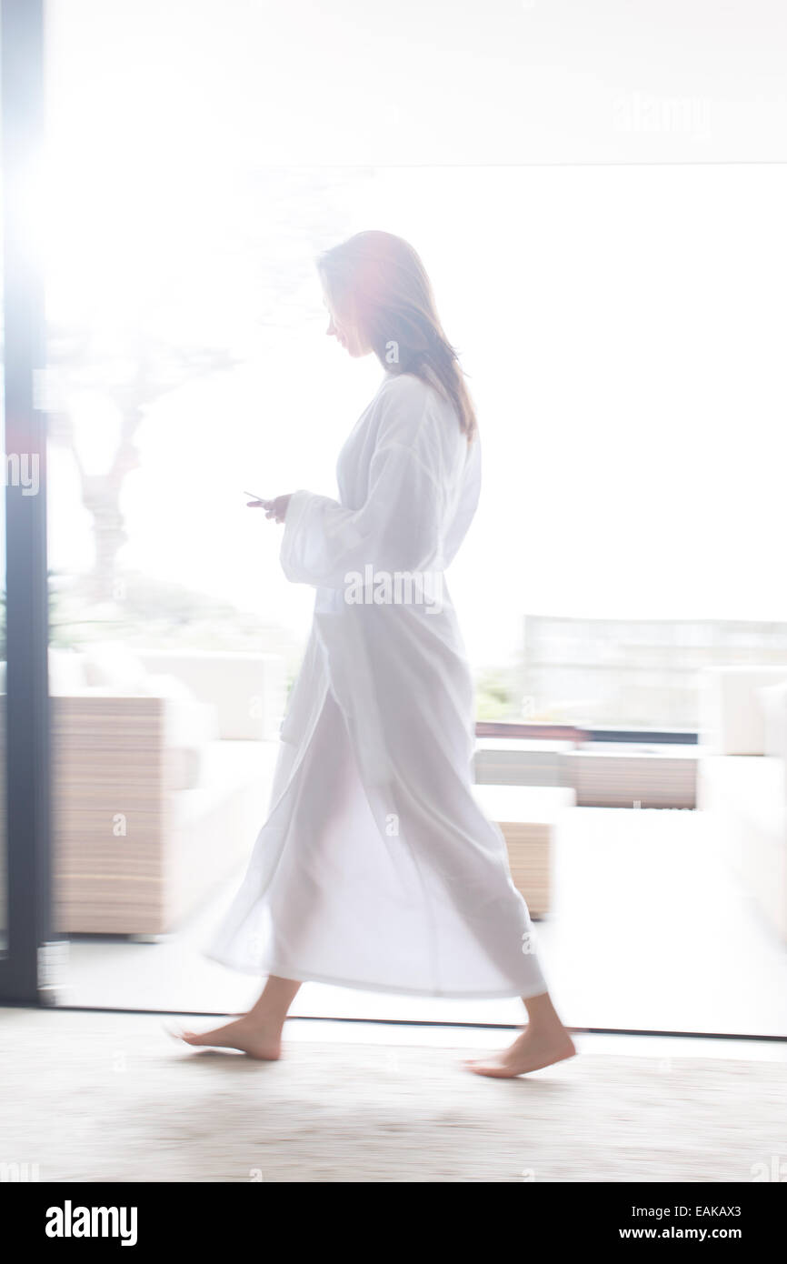 Woman wearing white bathrobe walking through modern corridor with mobile phone Stock Photo
