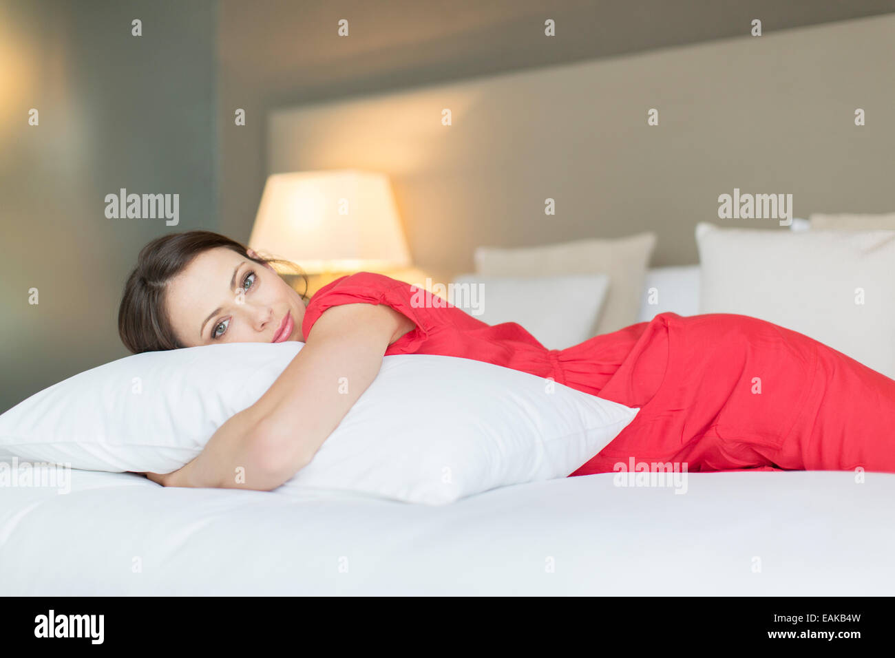 Portrait of beautiful woman wearing red dress lying on bed and hugging pillow Stock Photo