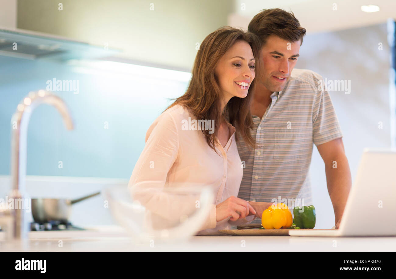 Smiling couple cutting bell peppers and looking at laptop in modern kitchen Stock Photo