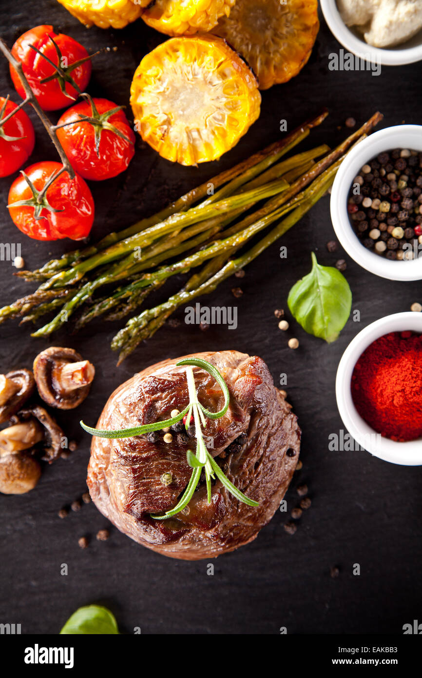 Piece of red meat steak with vegetable, herbs and spices, served on black stone surface. Shot from top view Stock Photo