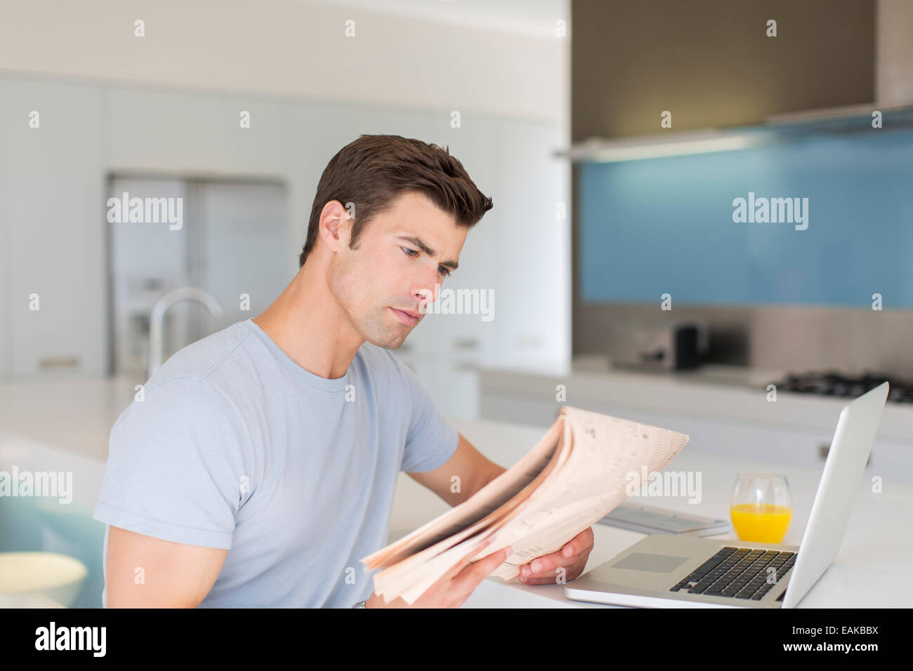 Man reading newspaper in modern kitchen, laptop and orange juice on counter Stock Photo