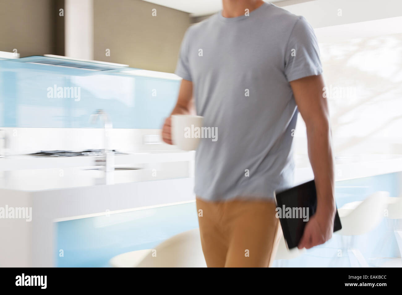 Man carrying coffee cup and digital tablet walking through modern kitchen Stock Photo