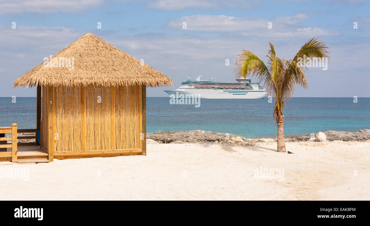 a bamboo cabana or hut with a grass roof situated on a sandy white beach along with a palm tree and cruise ship on the ocean. Stock Photo