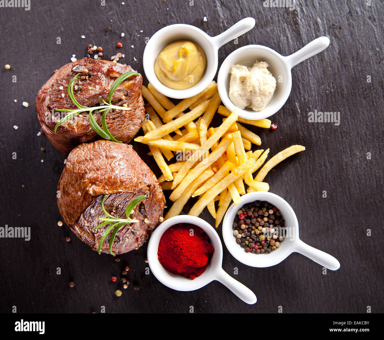 Pieces of red meat steaks with fries and spices, served on black stone surface. Stock Photo