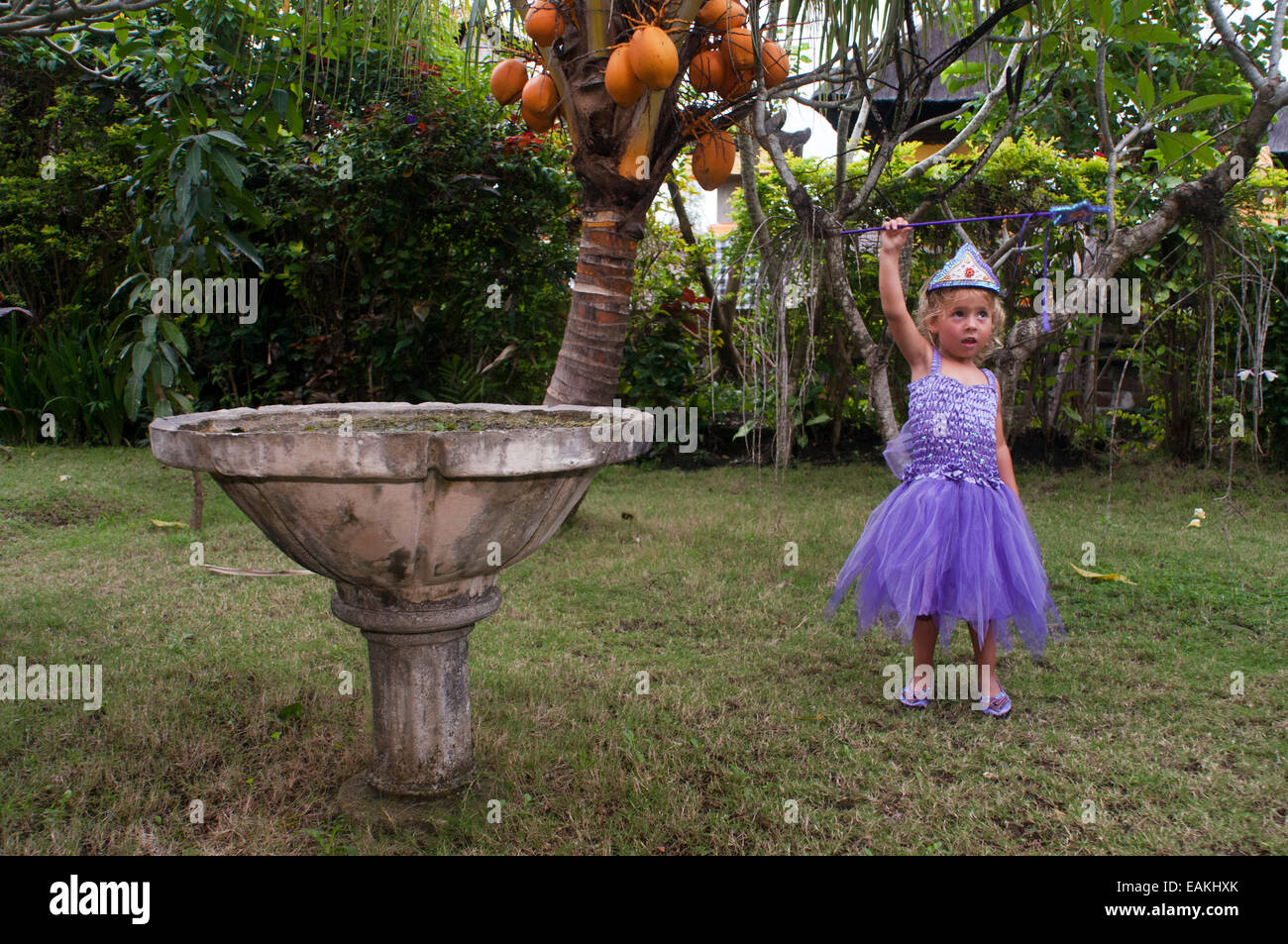 A girl dressed as princess with a magic wand in a Ubud garden. Indonesia. Party celebration. This stunning lilac dress has a bodice of lace, scattered with pink roses and sparkly sequins. Stock Photo