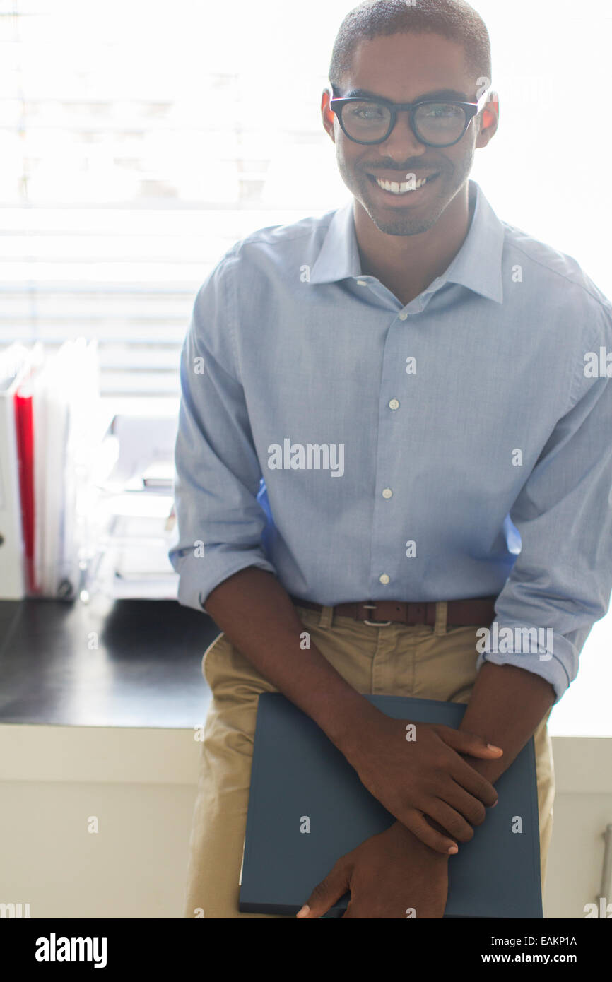 Portrait of smiling young man wearing glasses and blue shirt leaning on desk in office Stock Photo