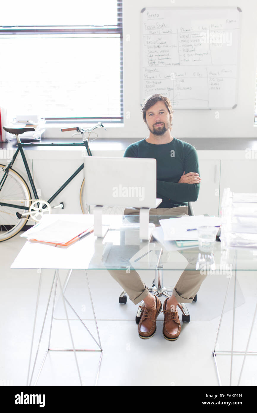 Portrait of man sitting with arms crossed behind glass desk in modern office, bicycle and whiteboard in background Stock Photo