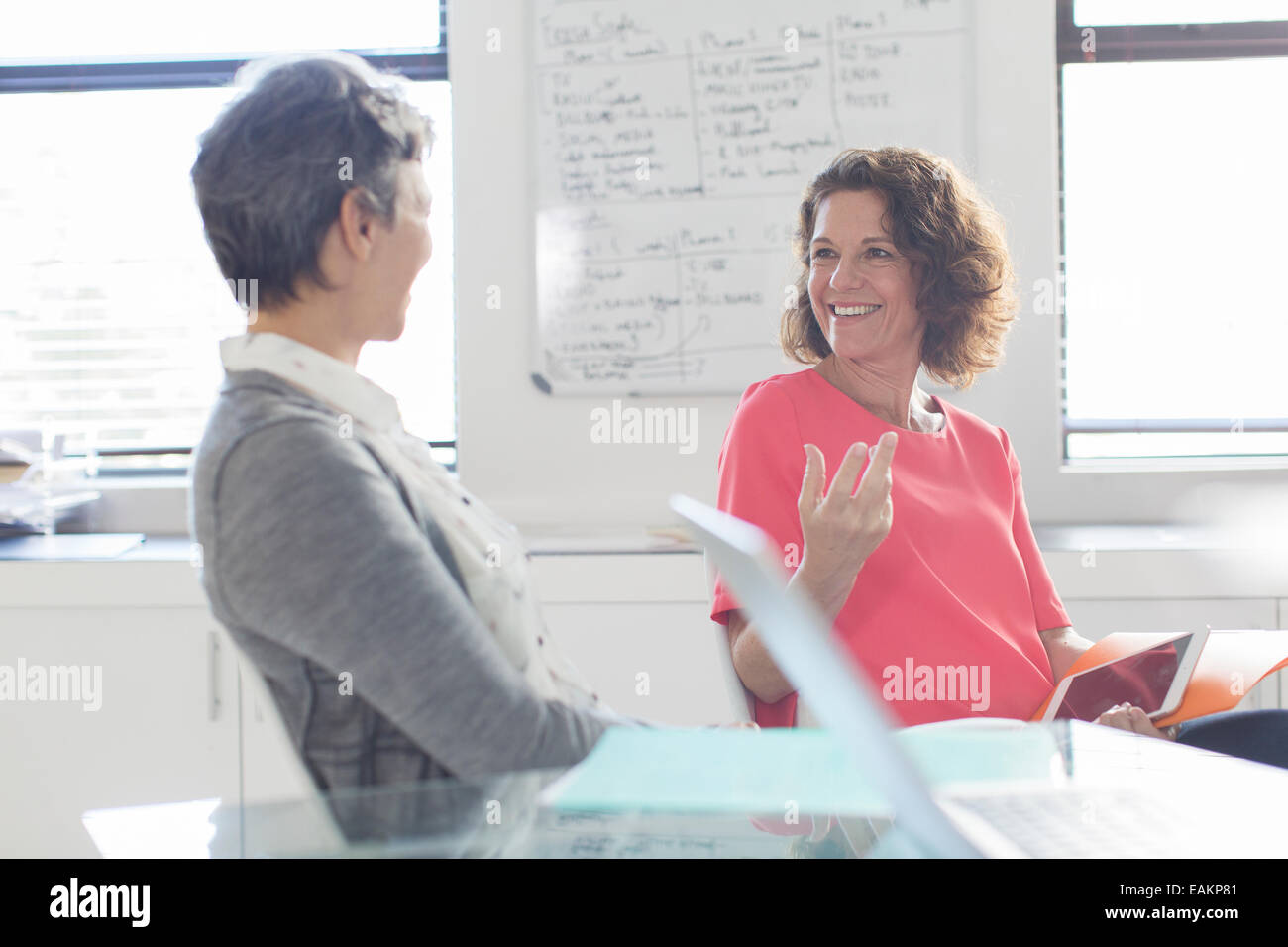 Two smiling women talking in office, whiteboard in background Stock Photo