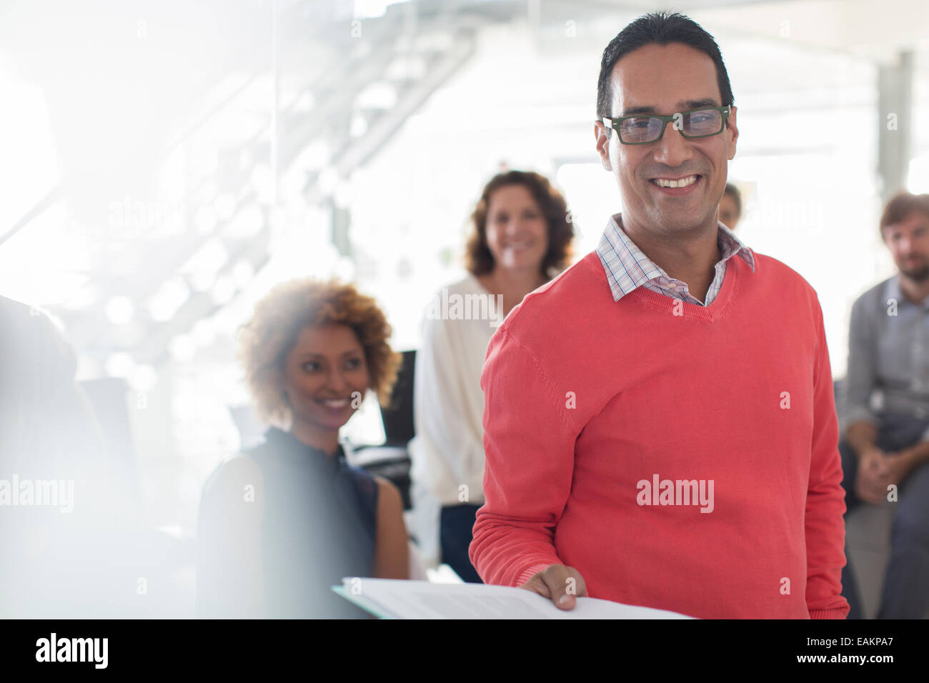 Portrait of smiling businessman wearing glasses and pink sweatshirt with team in background Stock Photo
