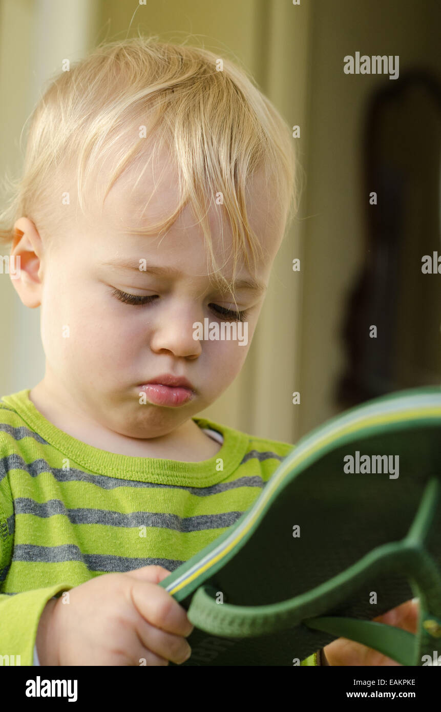 A blond and blue-eyed baby boy (ca. 18 months old) attentively studies a flip-flop. Stock Photo