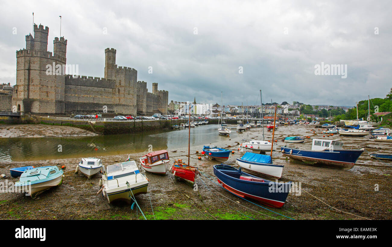 Huge, spectacular Caernarfon castle towering over River Seiont harbour & colourful boats with town in distance under stormy sky Stock Photo