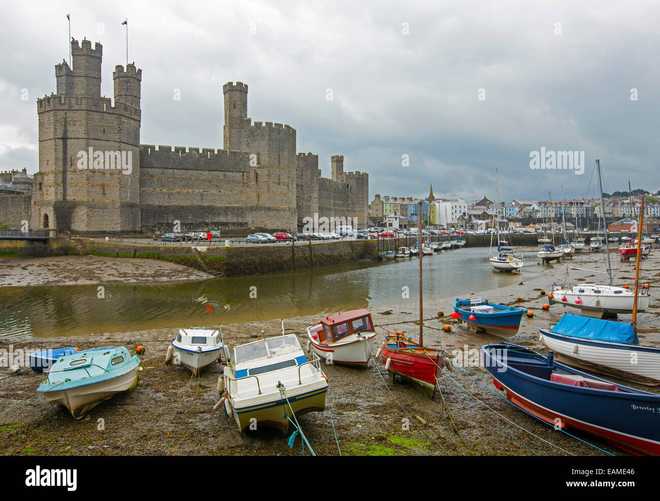 Huge, spectacular Caernarfon castle towering over River Seiont harbour & colourful boats with town in distance under stormy sky Stock Photo
