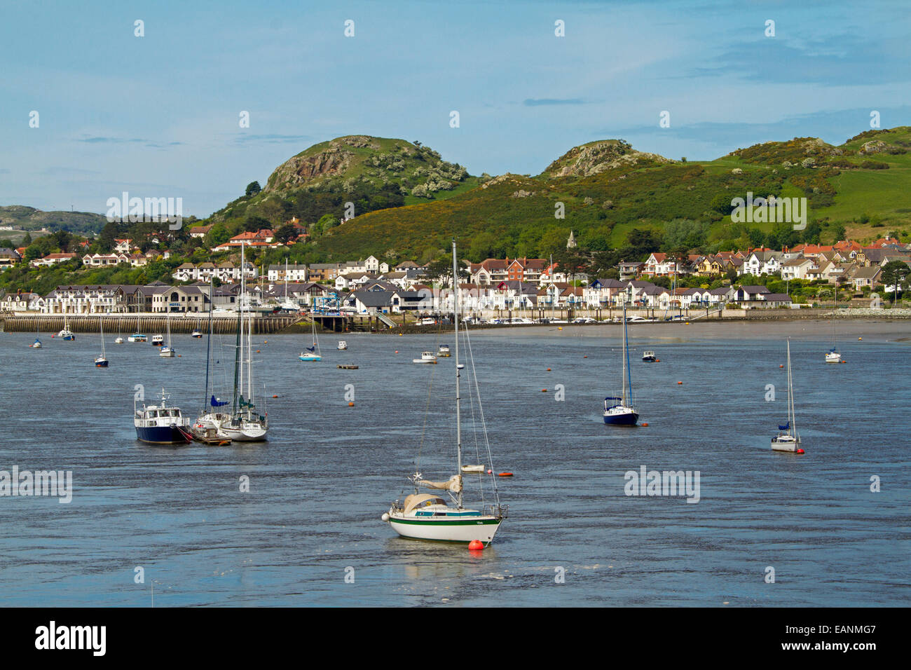 Boats moored on calm blue water of estuary of Conwy River with buildings of Welsh town of Conwy at base of adjacent hill Stock Photo