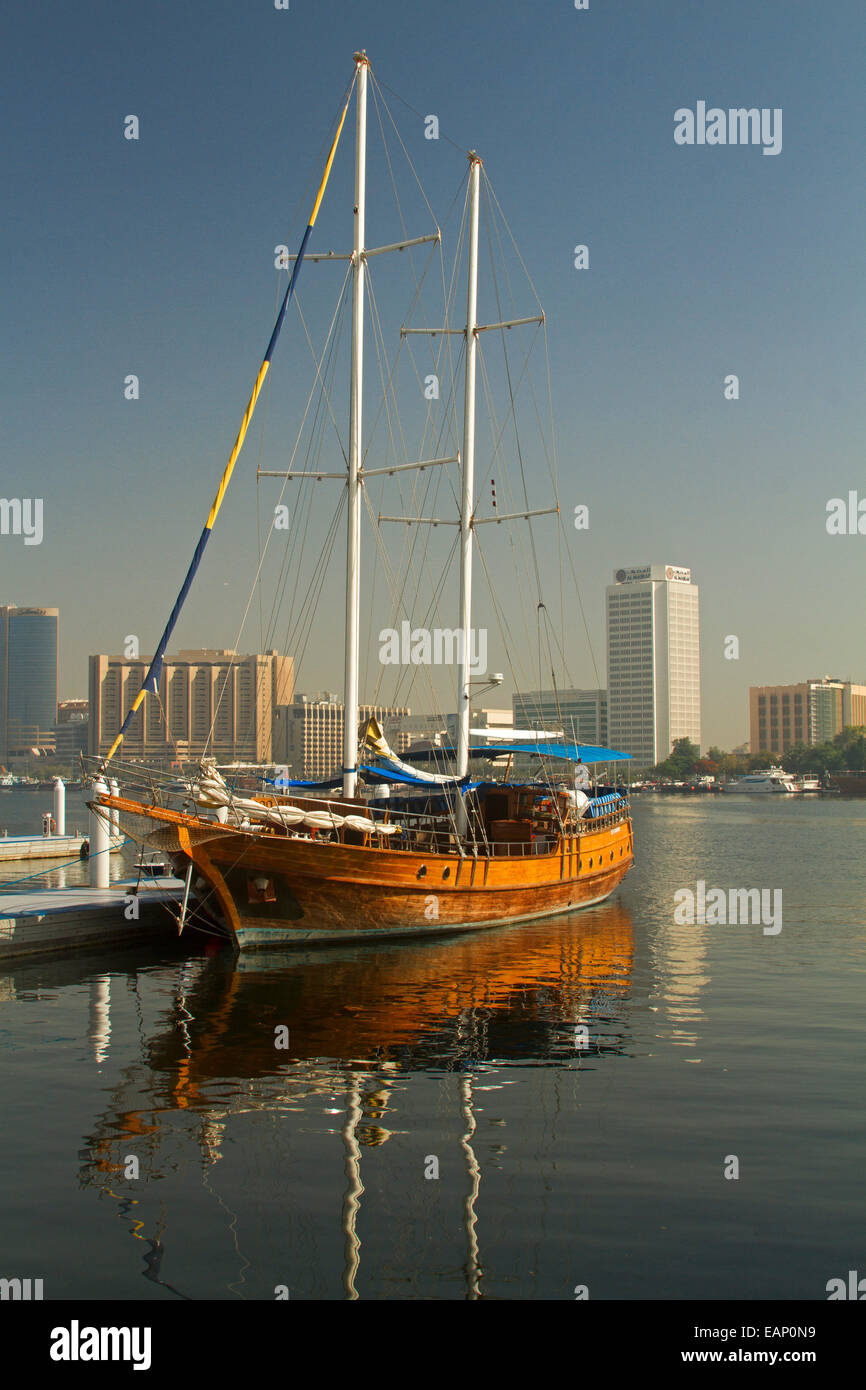 Wooden yacht moored & reflected in calm blue water of Dubai Creek with modern skyscrapers of city of Dubai in background under blue sky Stock Photo