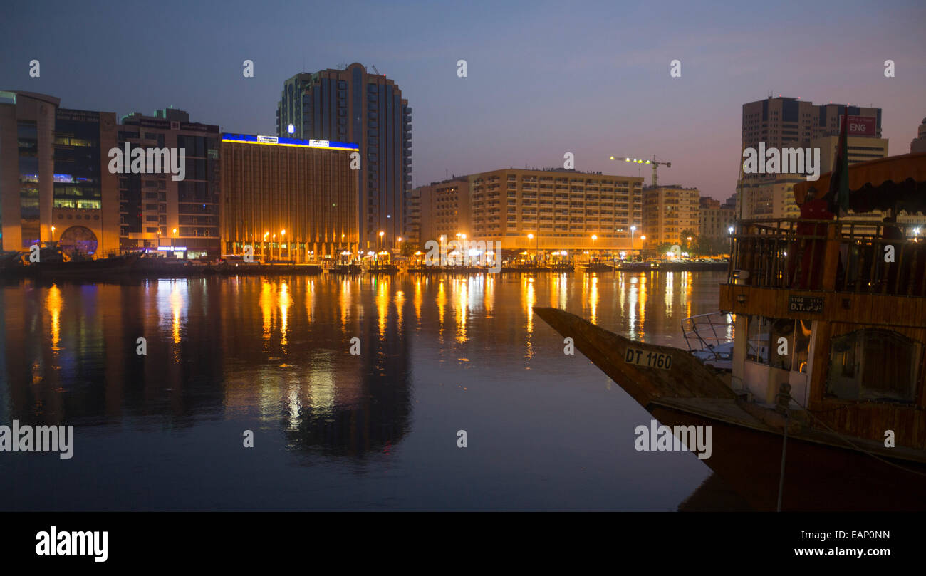 High-rise buildings against sky tinged with pink & bright lights of city reflected in dark calm waters of Dubai Creek at dawn Stock Photo