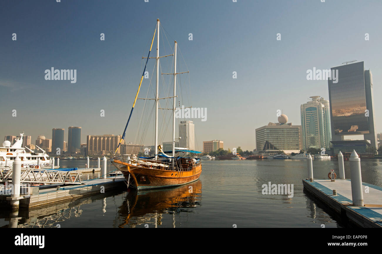 Wooden yacht moored & reflected in calm blue water of Dubai Creek with glass walled skyscraper & other modern buildings of city of Dubai nearby Stock Photo