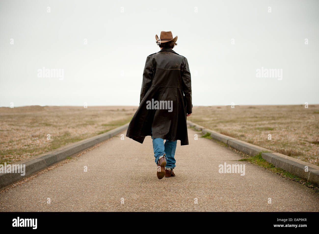 Man wearing long black and cowboy hat, walking away from camera towards the distant horizon. Stock Photo