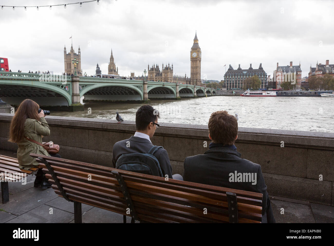 Two business men in suits and one female tourist sit on benches on next to the River Thames, opposite the Palace of Westminster Stock Photo