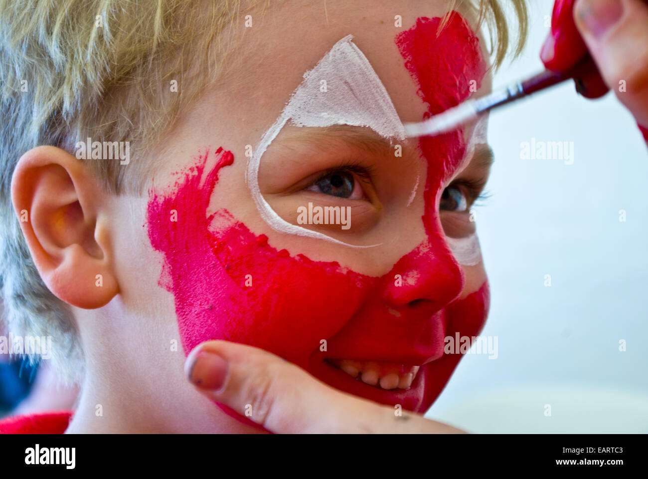 A blonde boy happily has his face painted bright red like spiderman. Stock Photo