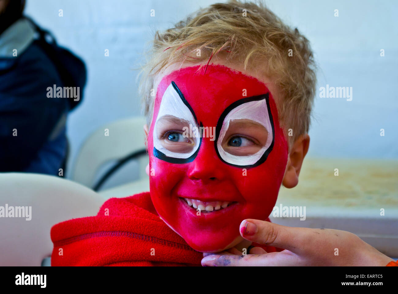 A blonde boy happily has his face painted bright red like spiderman. Stock Photo